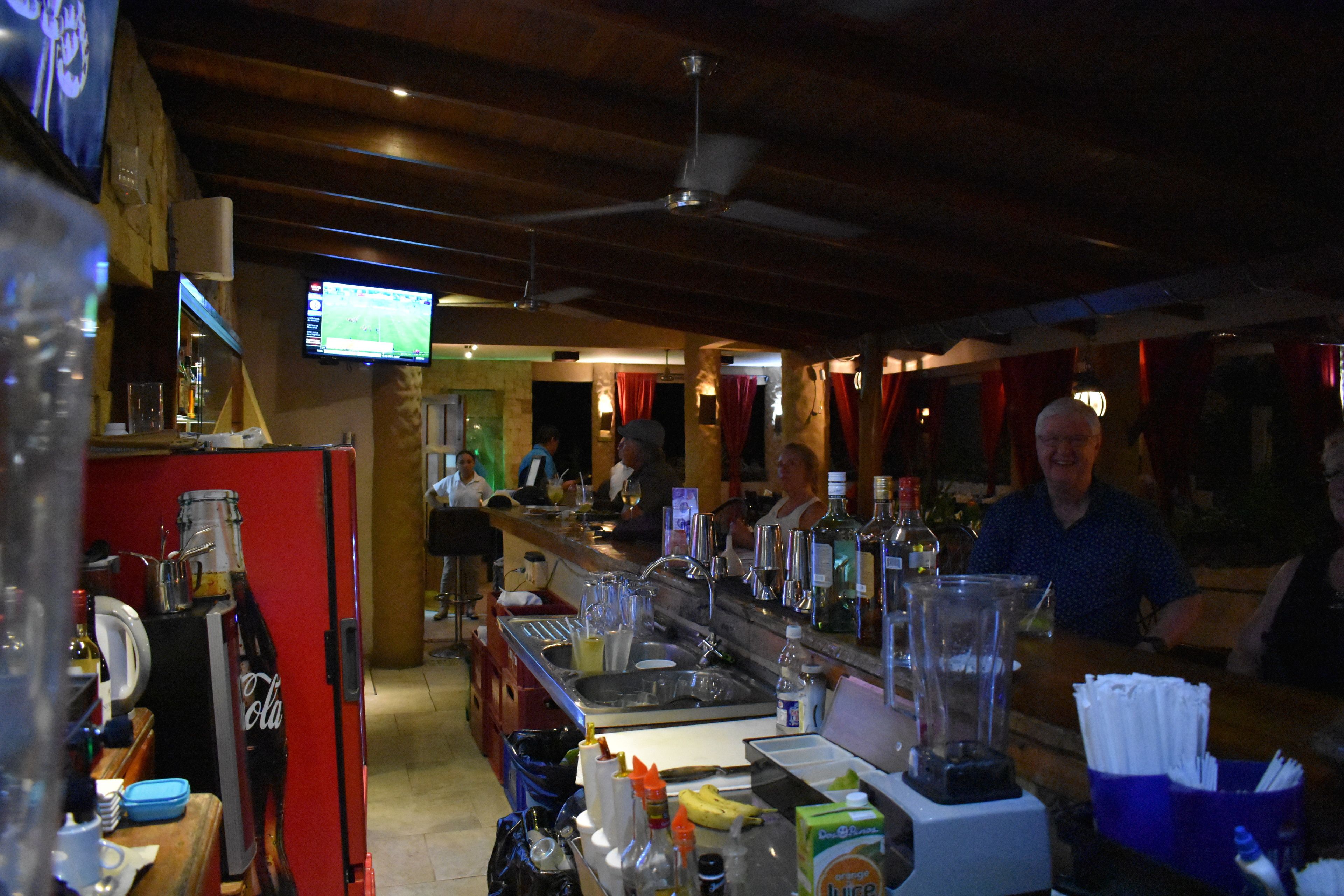 A bar area with a refrigerator, bottles, glasses, and a television displaying a sports game.