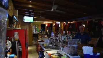 A bar area with a refrigerator, bottles, glasses, and a television displaying a sports game.