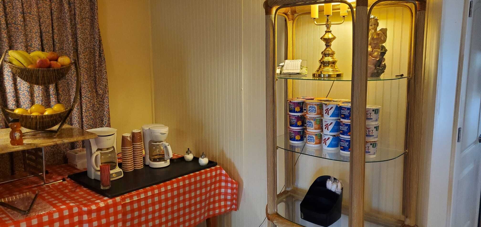 A room with a checkered tablecloth, a coffee station, a fruit basket, and a glass cabinet with canned goods.