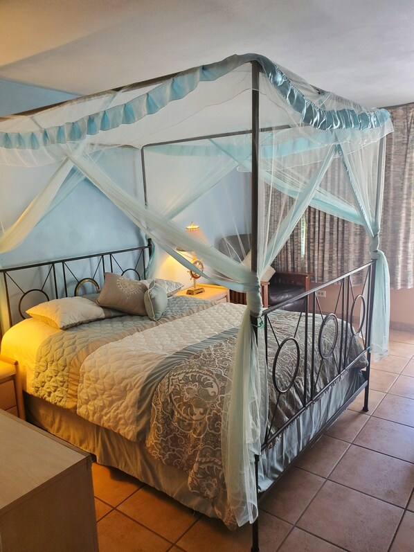 A four-poster bed with a canopy, a bedside table with a lamp, and a view of a room with a window and curtains.