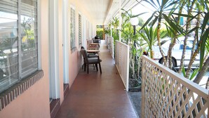 A covered corridor with tables and chairs, palm trees, and a vehicle outside.