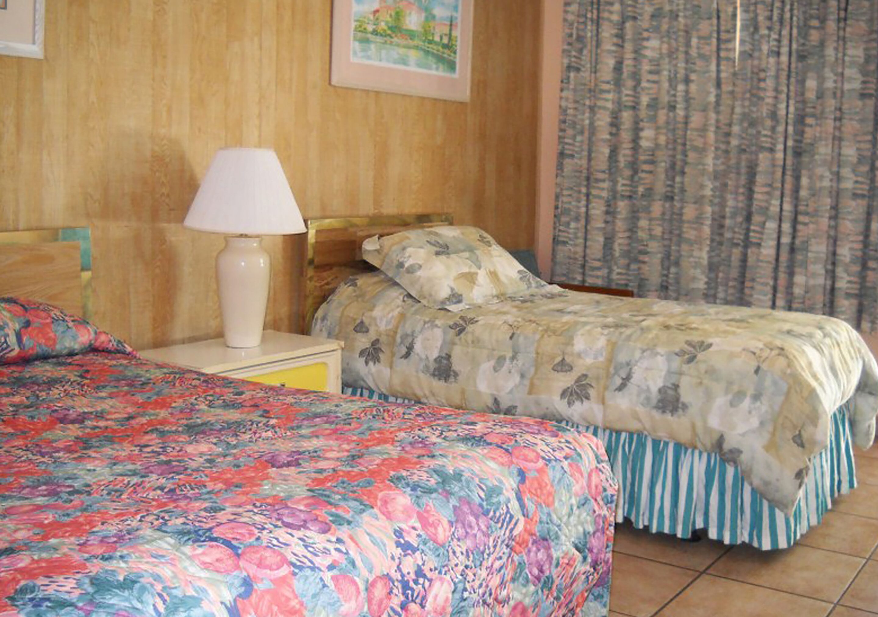 Habitación de hotel con dos camas, una mesita de noche con lámpara, cabecera de madera y un cuadro en la pared.