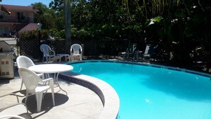 A swimming pool with a tiled edge, surrounded by white plastic chairs and a table.