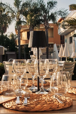 A table set with wine glasses, forks, and a woven placemat outdoors, with a lamp and umbrellas in the background. - Hotel Première Pool & Resort (Giugliano in Campania)