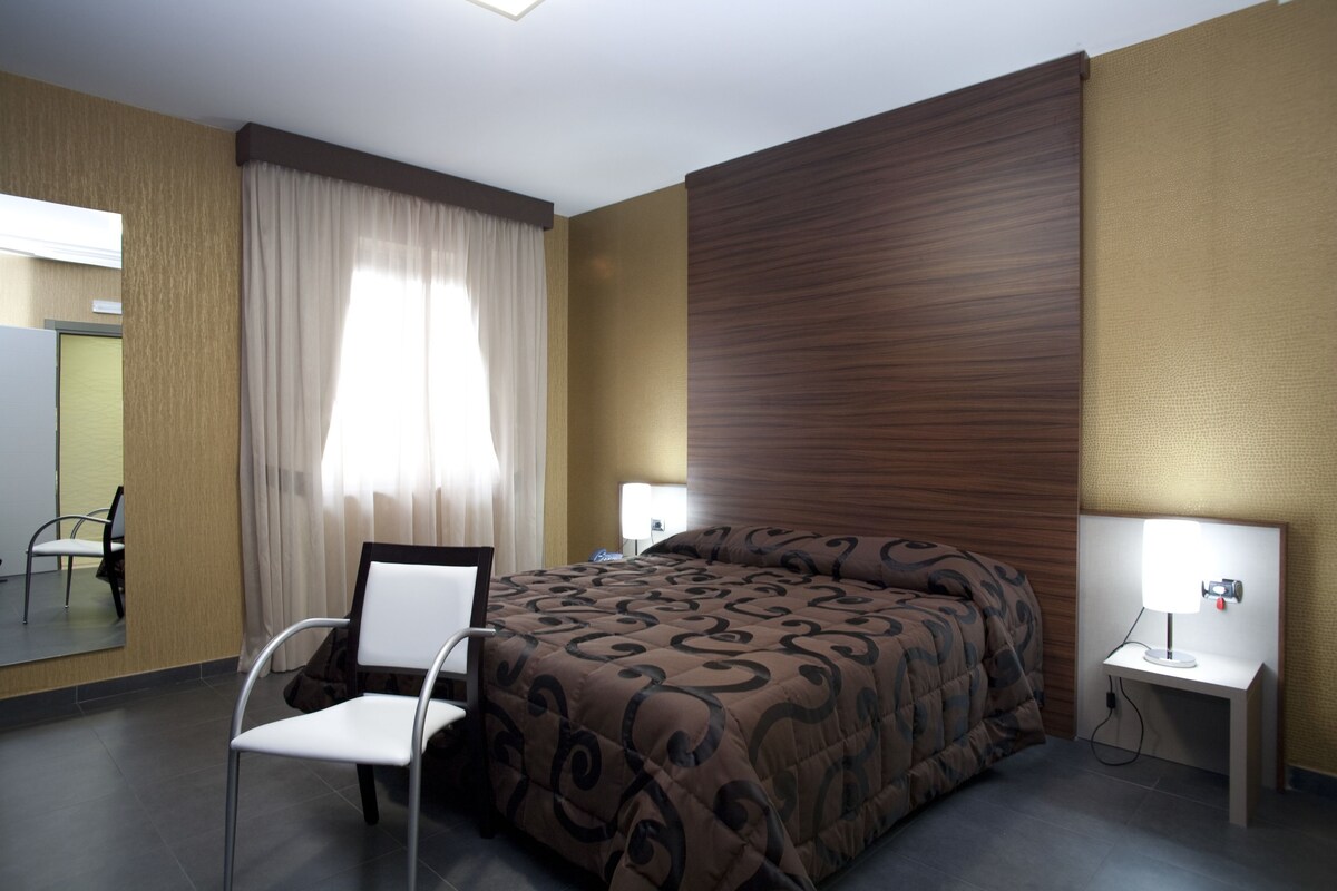 A hotel room with a bed, a chair, a desk with a lamp, and a mirror.