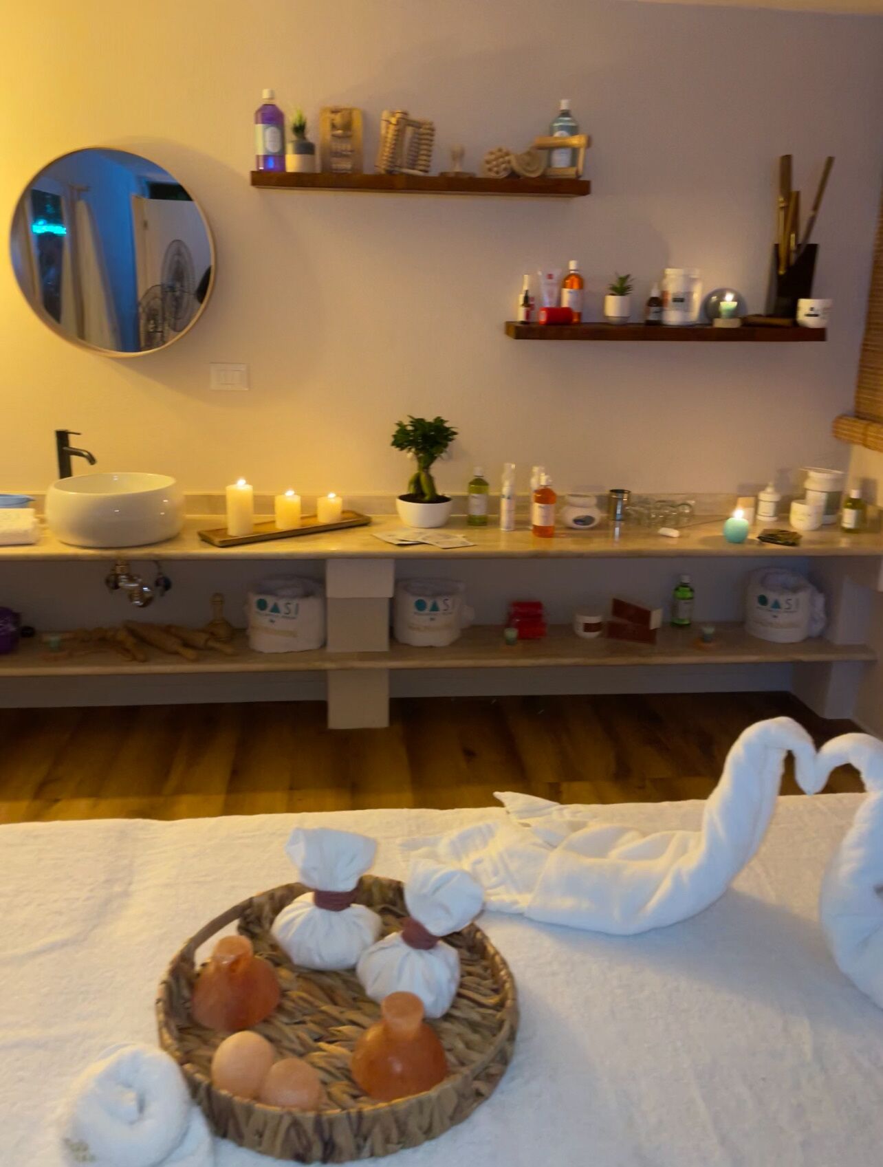 A bathroom with a round mirror, a sink, shelves with toiletries, and a decorative towel arrangement.