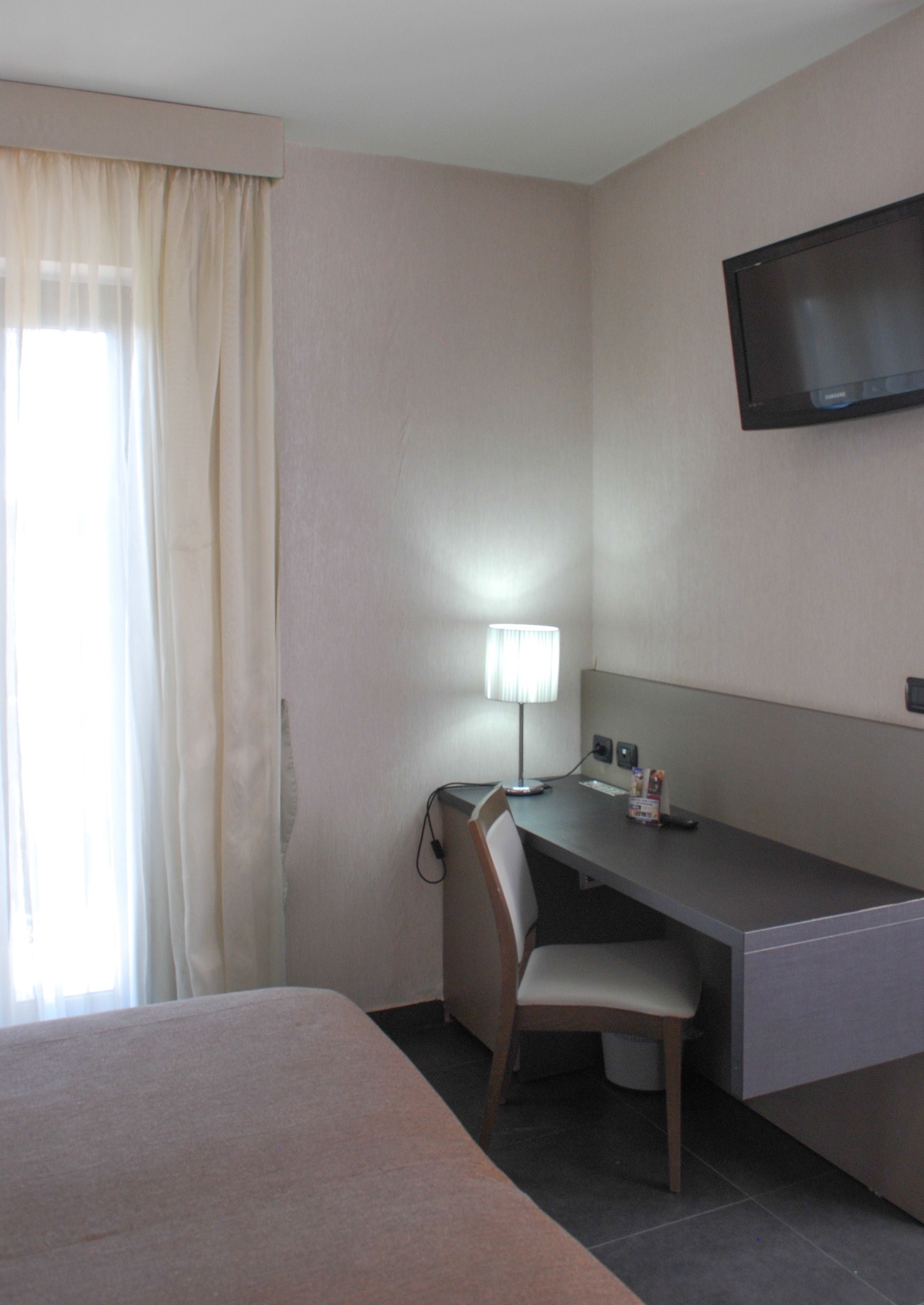 Habitació d'hotel amb llit, escriptori amb cadira, làmpada, televisió i finestra amb cortines.