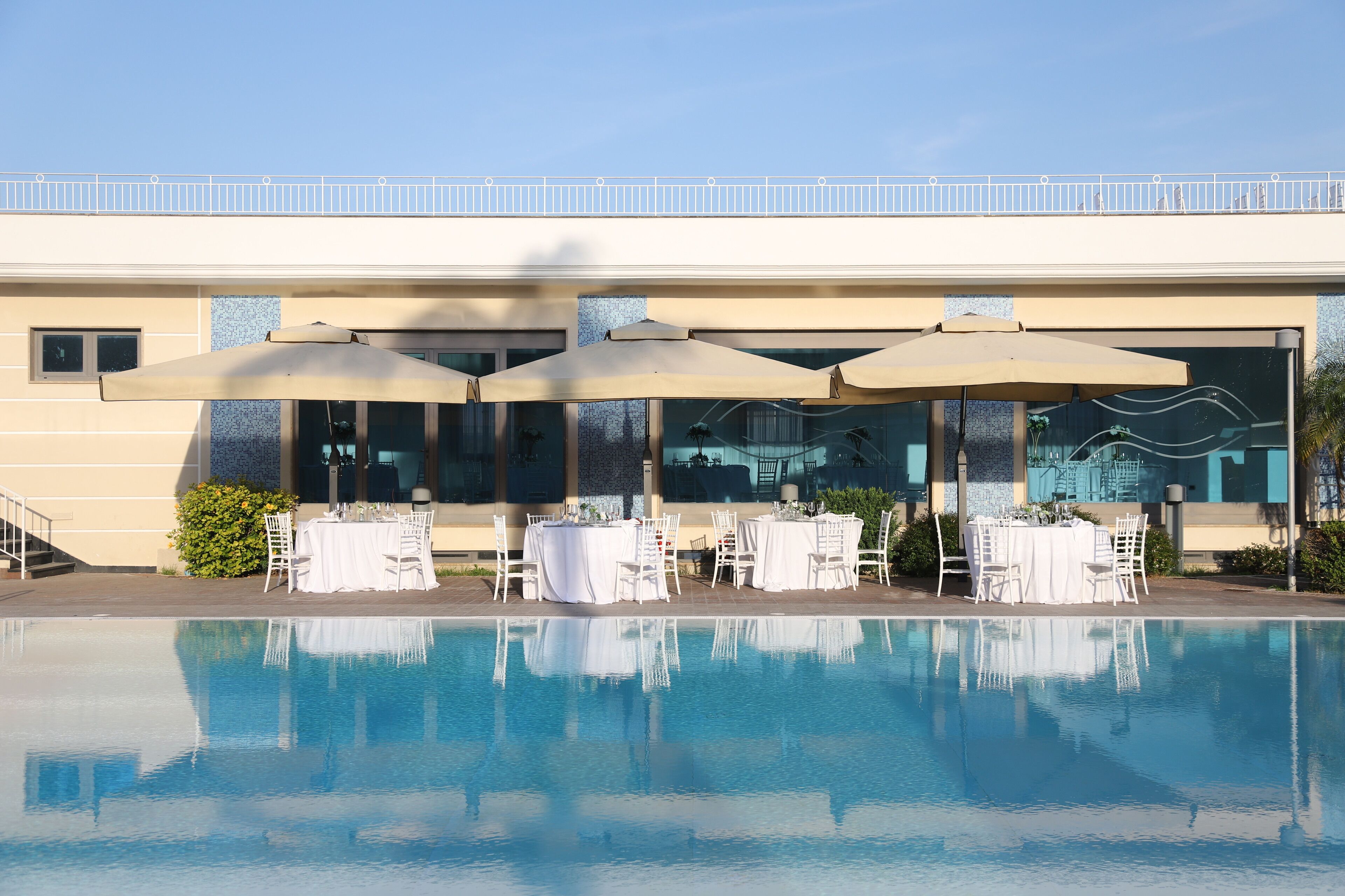 Un área junto a la piscina con mesas y sillas, piscina cristalina y un edificio con balcón al fondo.