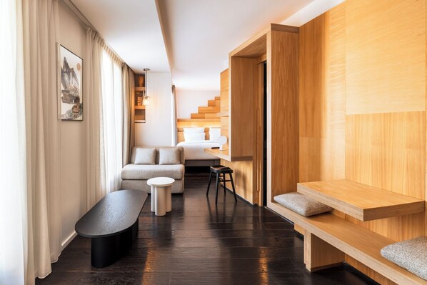 A modern hotel room with a sofa, a small round table, a bench, and a bed with a wooden headboard.
