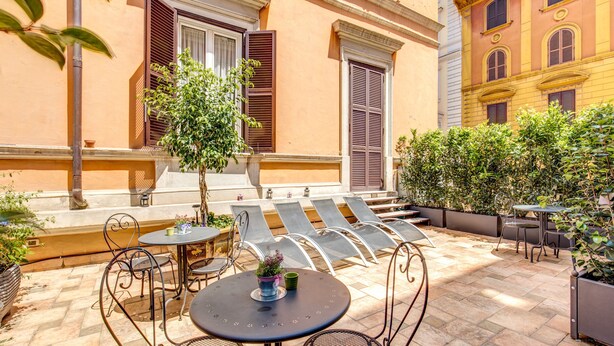 An outdoor seating area with metal chairs and tables, surrounded by potted plants and a building with shuttered windows. - Roman Terrace (Rome)