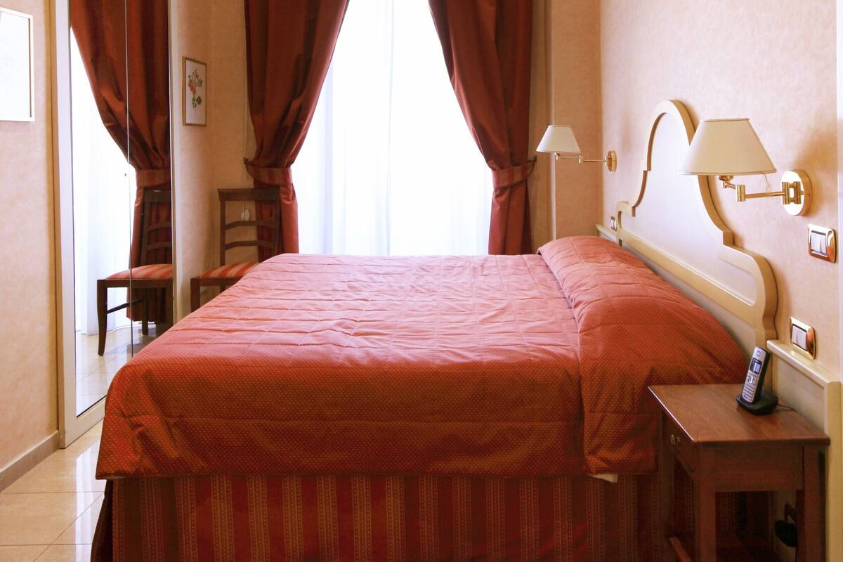 Una camera d'albergo con un letto grande, un comodino, una lampada e una finestra con tende.