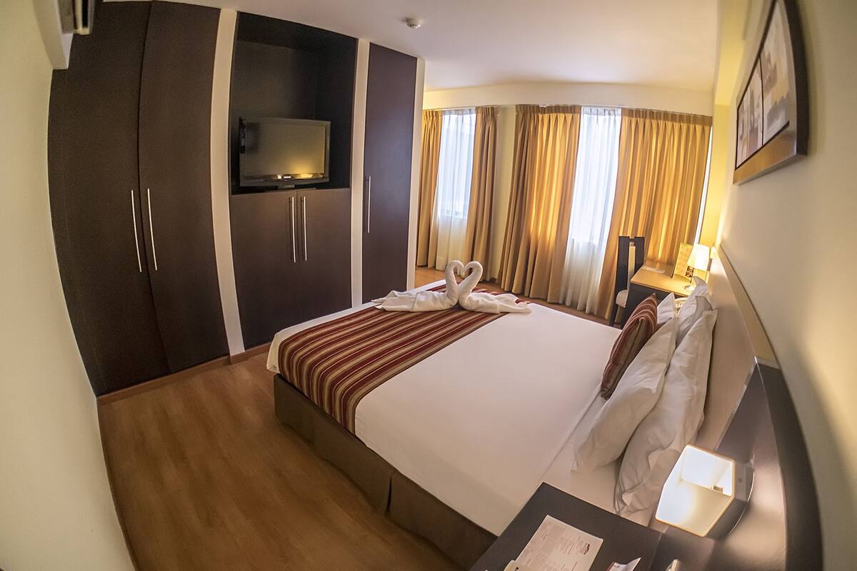 A hotel room with a large bed, a TV on a stand, a desk with a lamp, and a wardrobe.