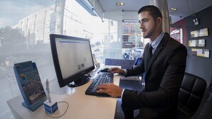 A man working at a desk with a computer, a keyboard, and a mouse. There is a window behind him, and a sign for a hotel is visible outside. - Allpa Hotel & Suites (Lima)