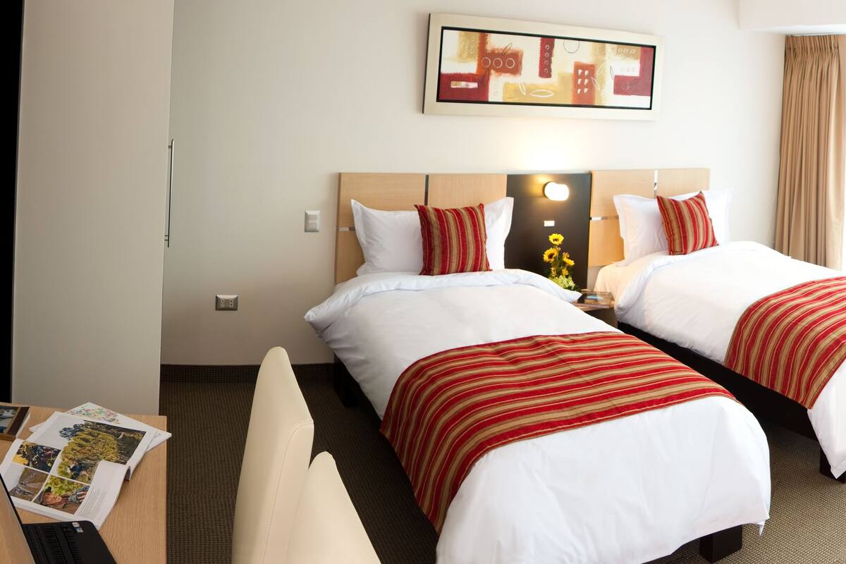 A hotel room with two beds, a desk, and a chair. There is a framed picture on the wall and a window with curtains.