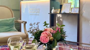 A reception area with a sign for Chateau d'Apigne Hotel & Gastronomie, a reception desk, a chair with a cushion, a glass table with drinks and a vase of flowers.
