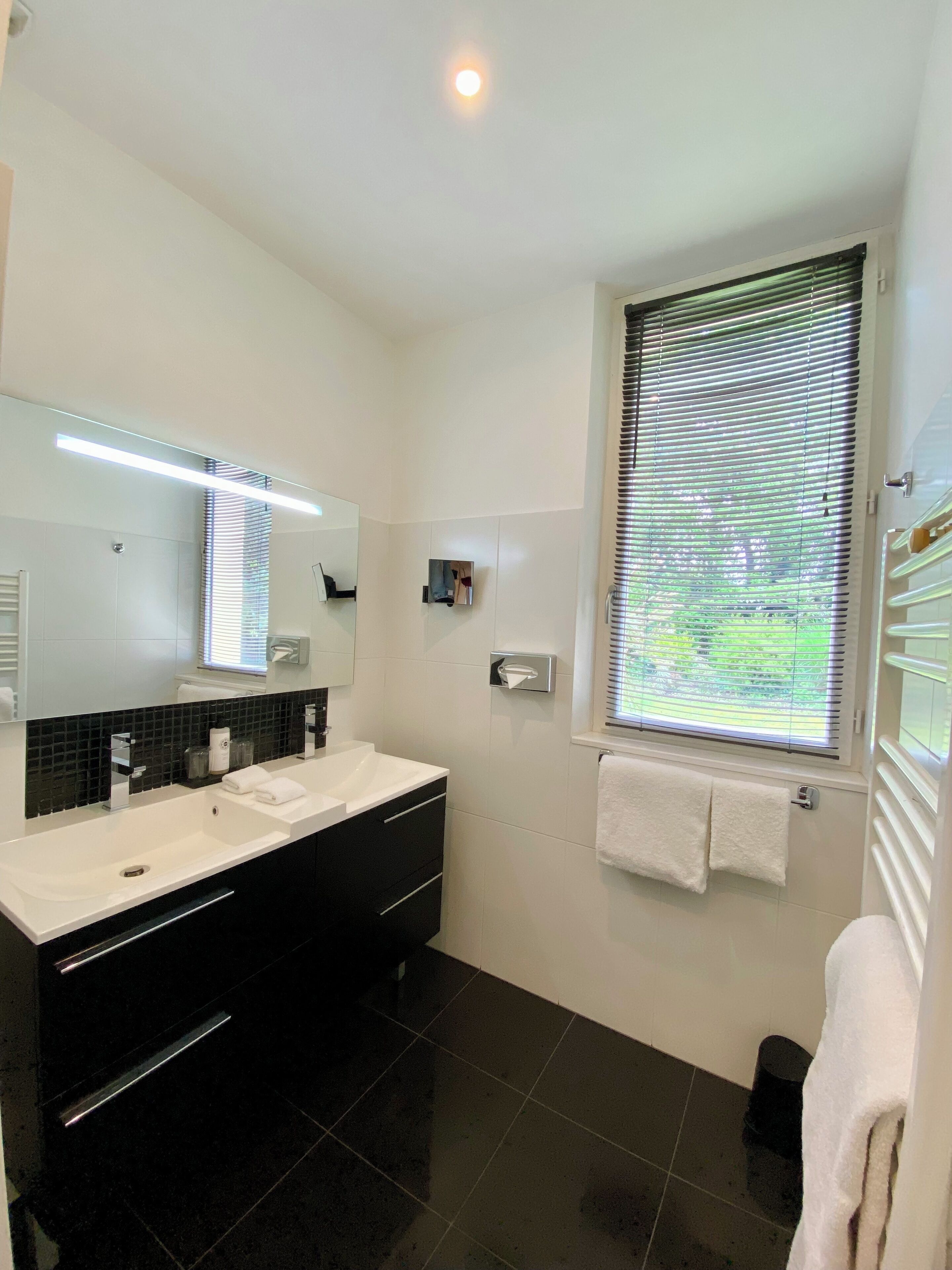A modern bathroom with a large mirror, two sinks, black cabinets, and a window with blinds.