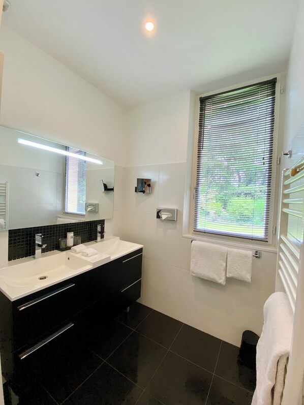 A modern bathroom with a large mirror, two sinks, black cabinets, and a window with blinds.