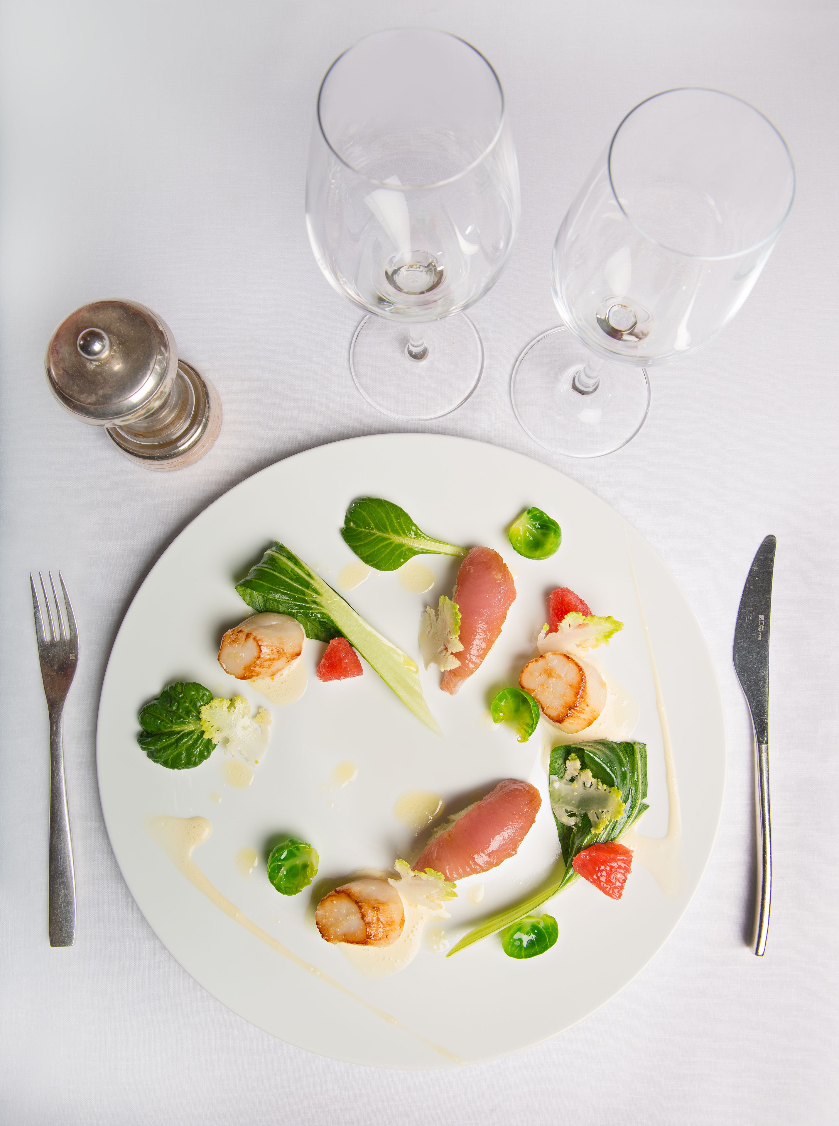 A white plate with a seafood dish, two empty wine glasses, a salt shaker, and a fork and knife on a white table.