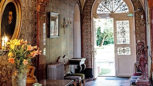 An ornate hallway with a chandelier, a framed portrait, and a large vase of flowers.