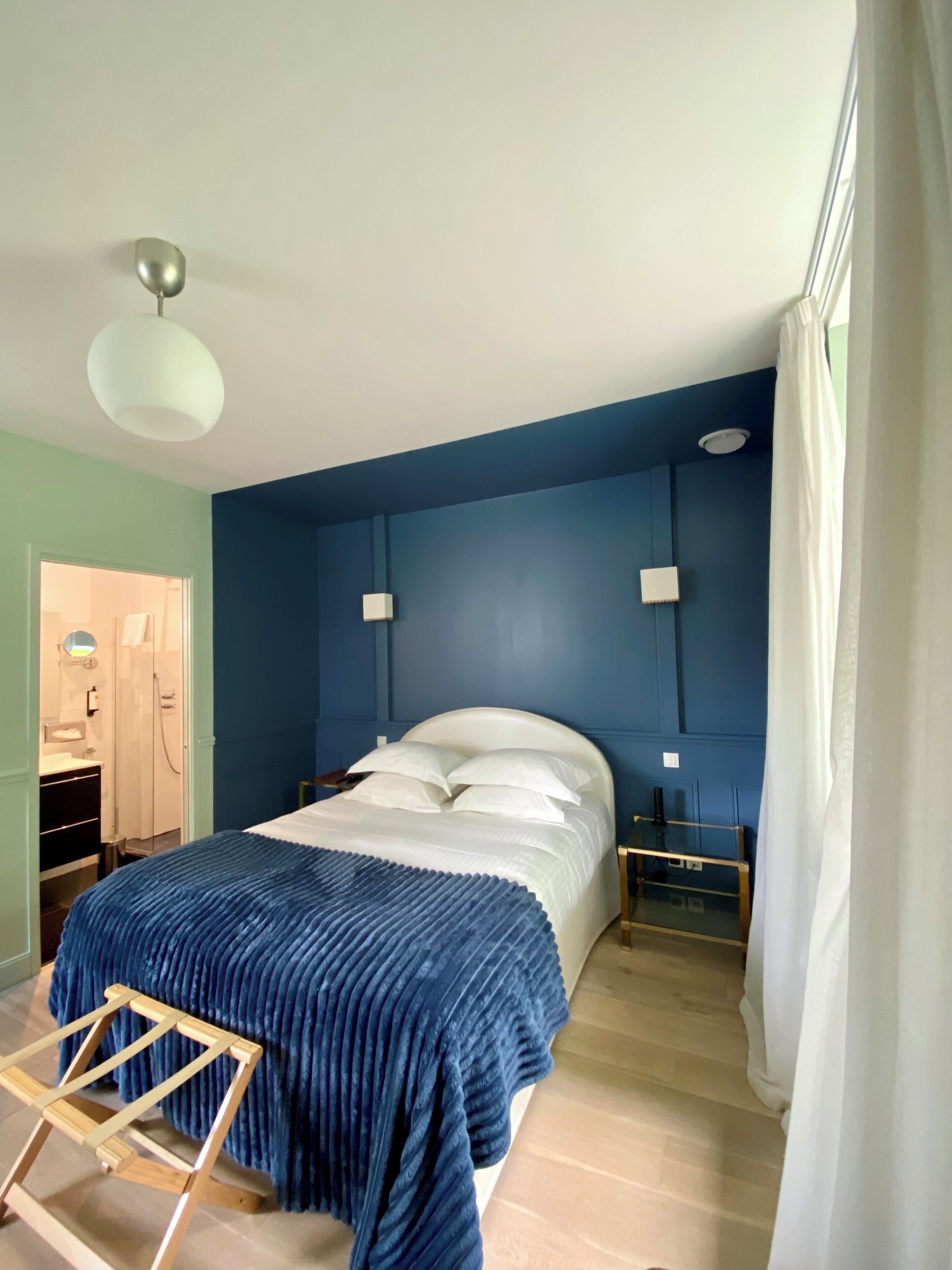 Une chambre à coucher avec un lit, des tables de chevet, une chaise en bois, un miroir et une fenêtre avec des rideaux.