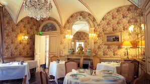 A dining area with tables set for service, a chandelier, floral wallpaper, and a mirror with a painting.