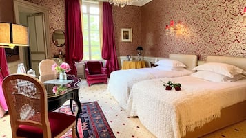 A hotel room with two beds, a chandelier, a dining table with chairs, a patterned rug, and a large mirror.