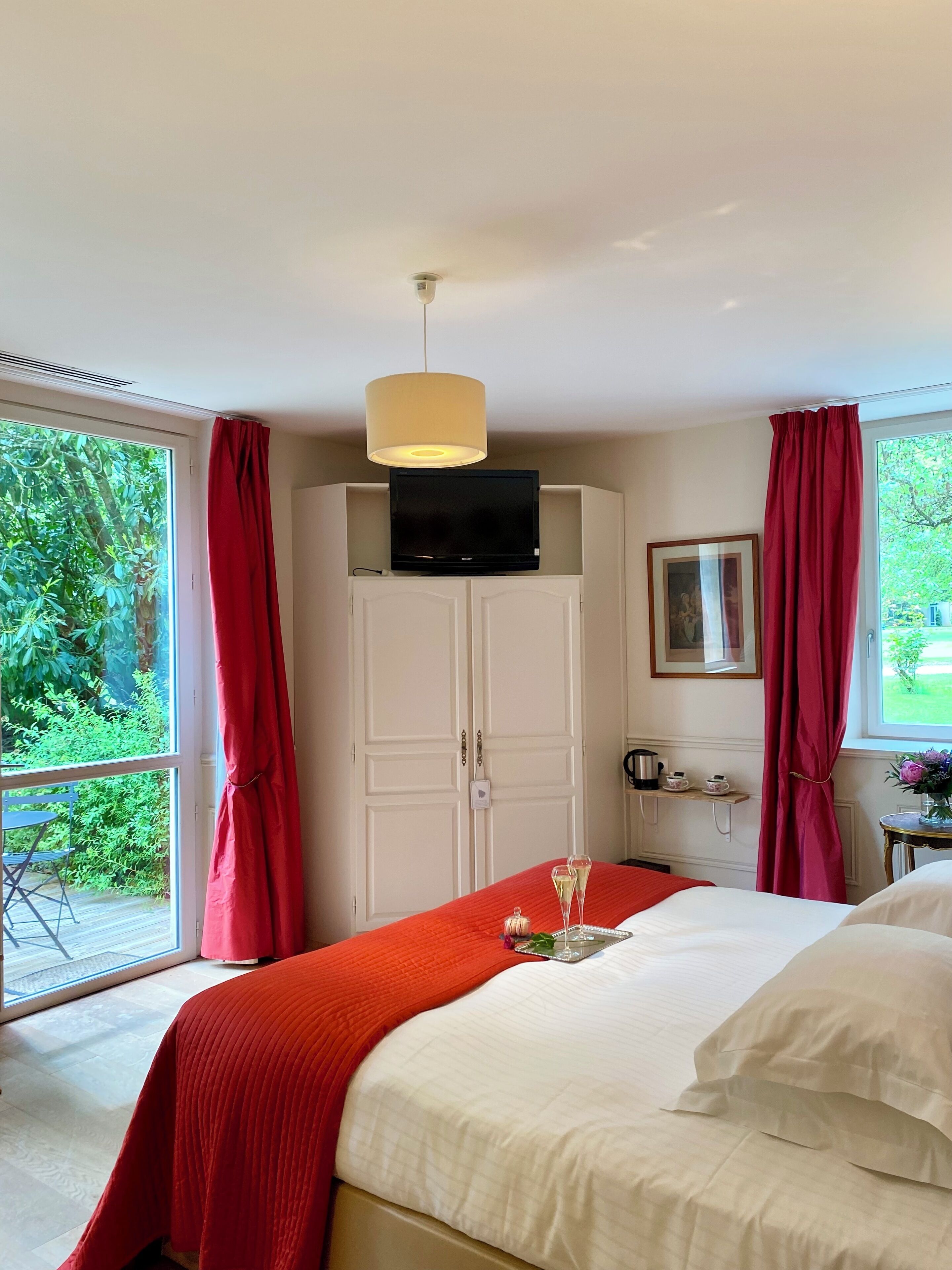 Un dormitorio con cama, televisión sobre un mueble, ventana con cortinas, una mesita con un florero y vistas al exterior con vegetación.