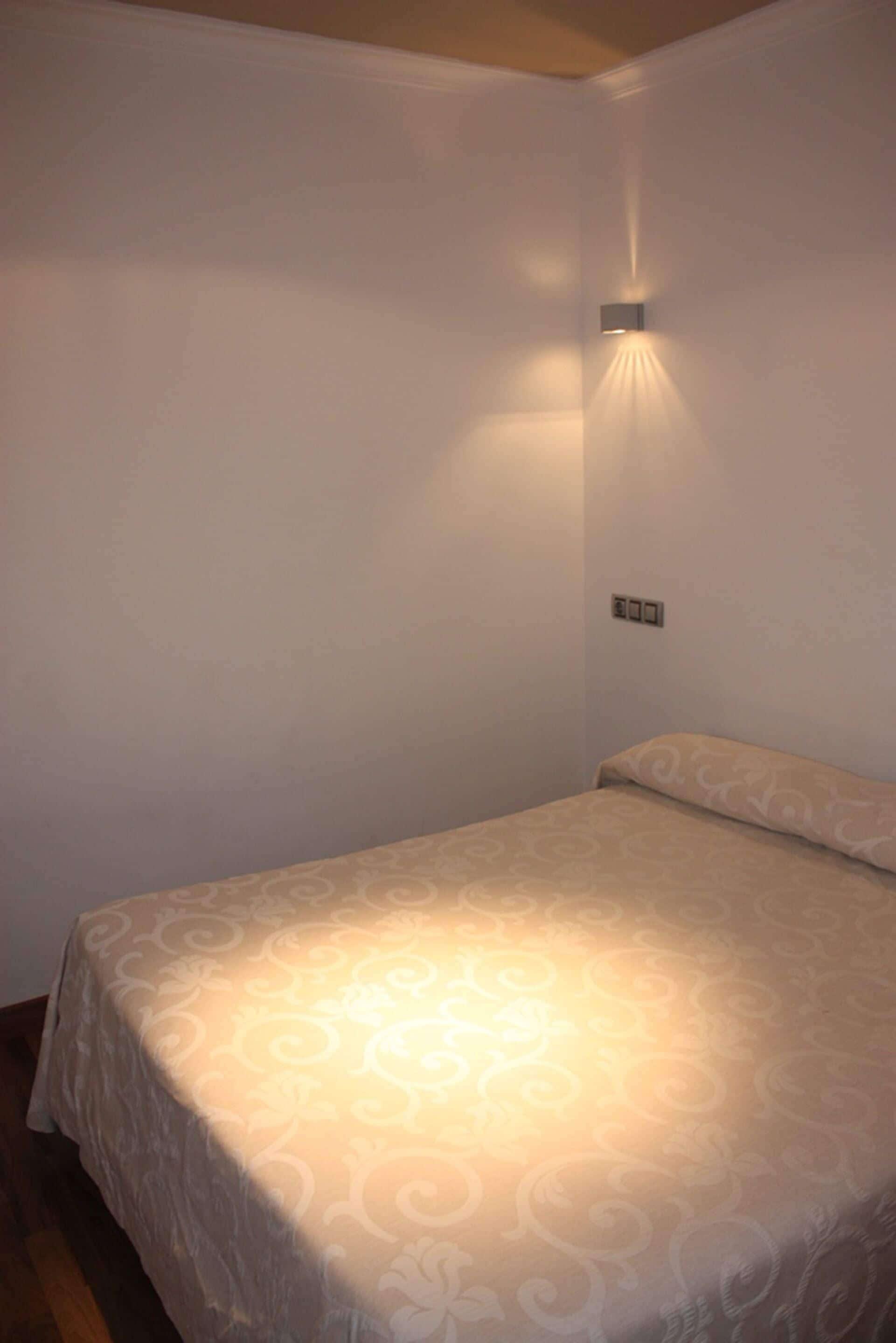 A single bed with a patterned bedspread in a room with white walls and a wall-mounted light fixture.
