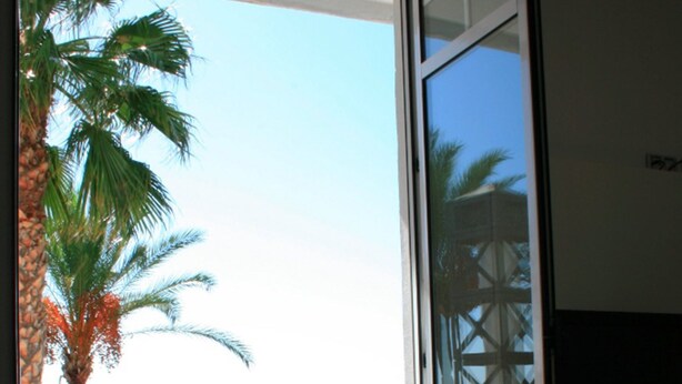Un balcón con puerta de vidrio, una palmera y vista a una playa con sombrillas y gente.