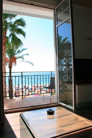 A balcony with a glass door, a palm tree, and a view of a beach with umbrellas and people. - Apartamentos Senabre Palais (Benidorm)