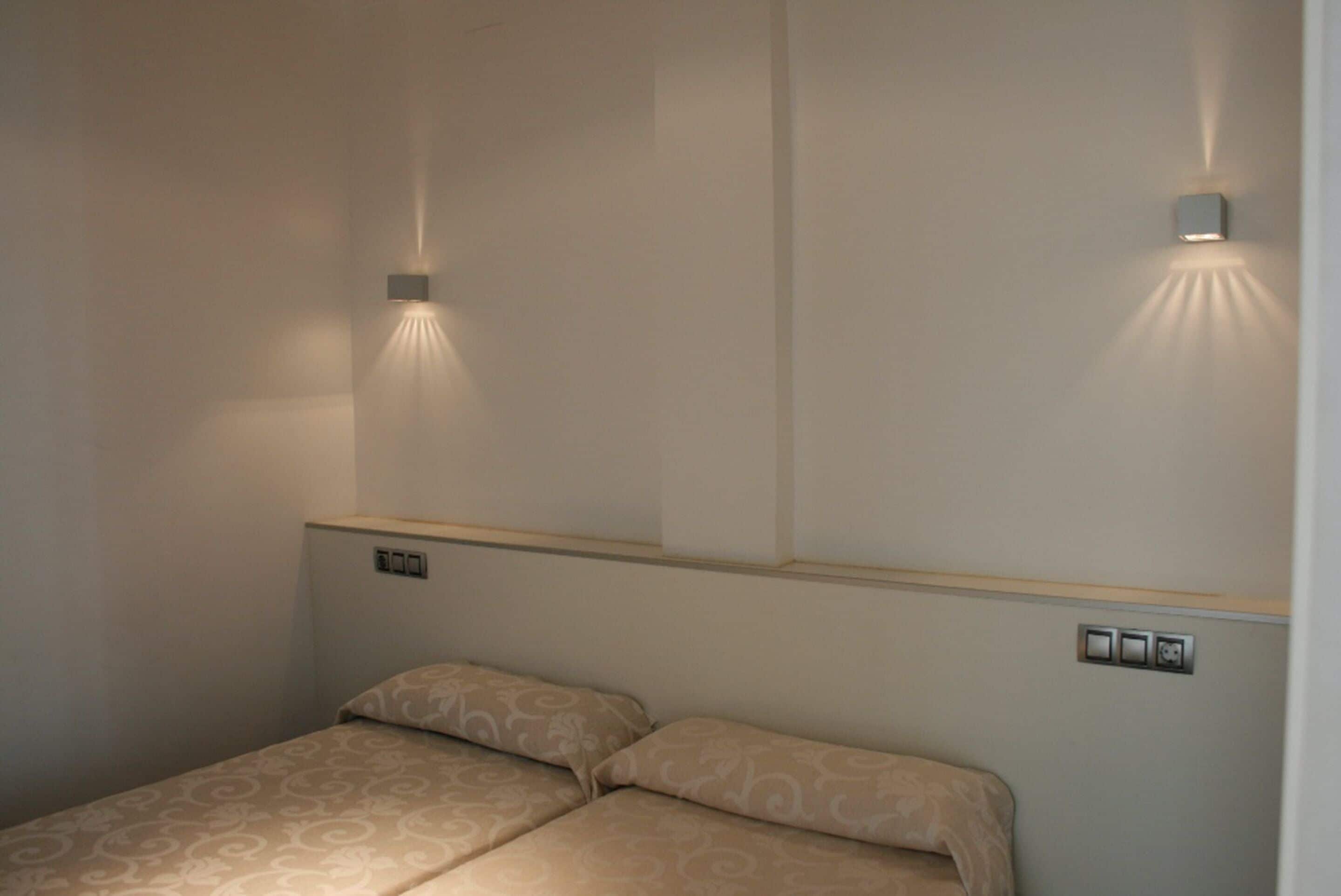 Una habitación de hotel con una cama, dos lámparas fijadas a la pared y un pequeño panel de control.