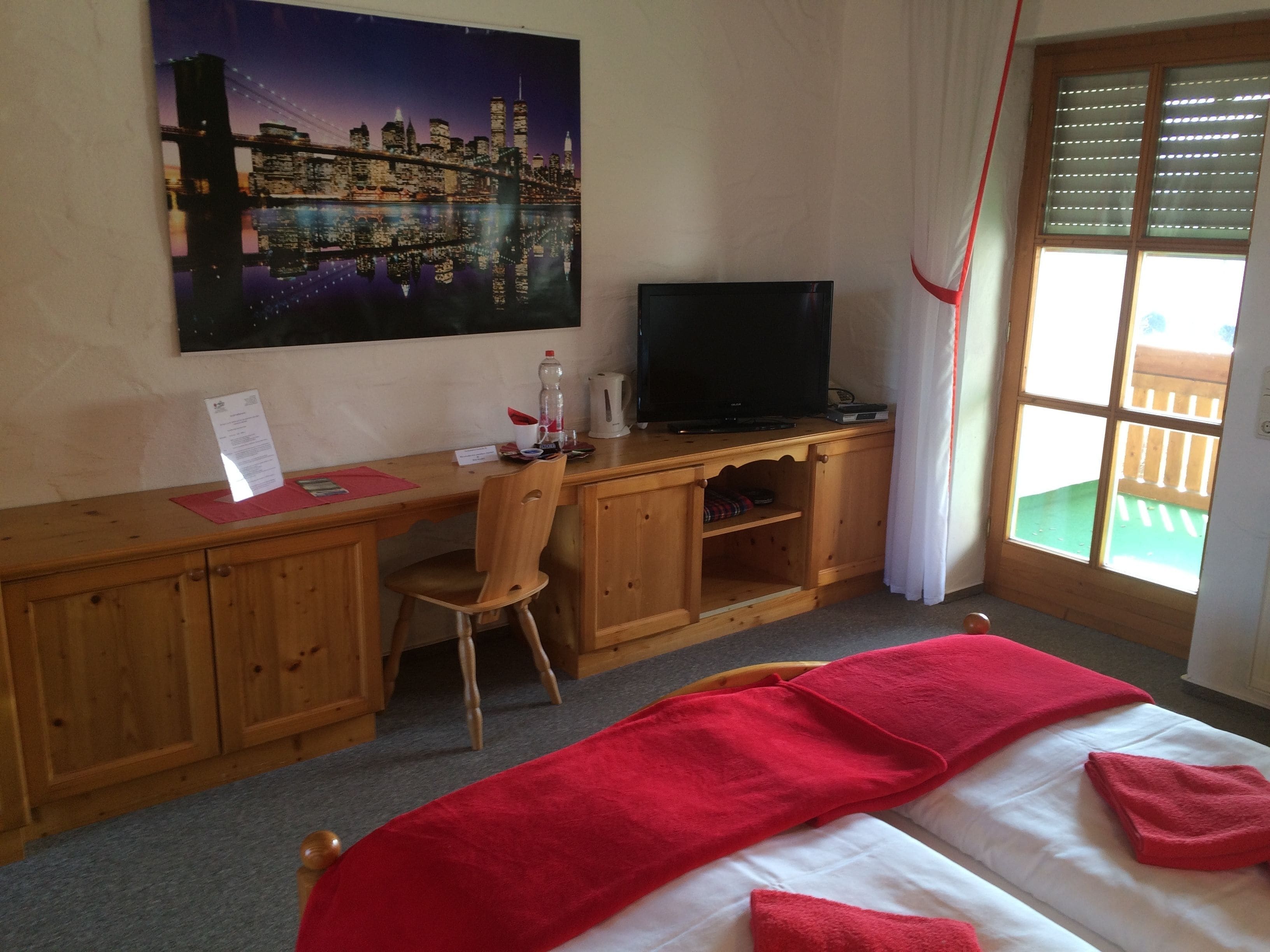 A hotel room with a wooden desk, a television, a bed with red towels, a cityscape painting, and a window with blinds.