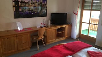 A hotel room with a wooden desk, a television, a bed with red towels, a cityscape painting, and a window with blinds.