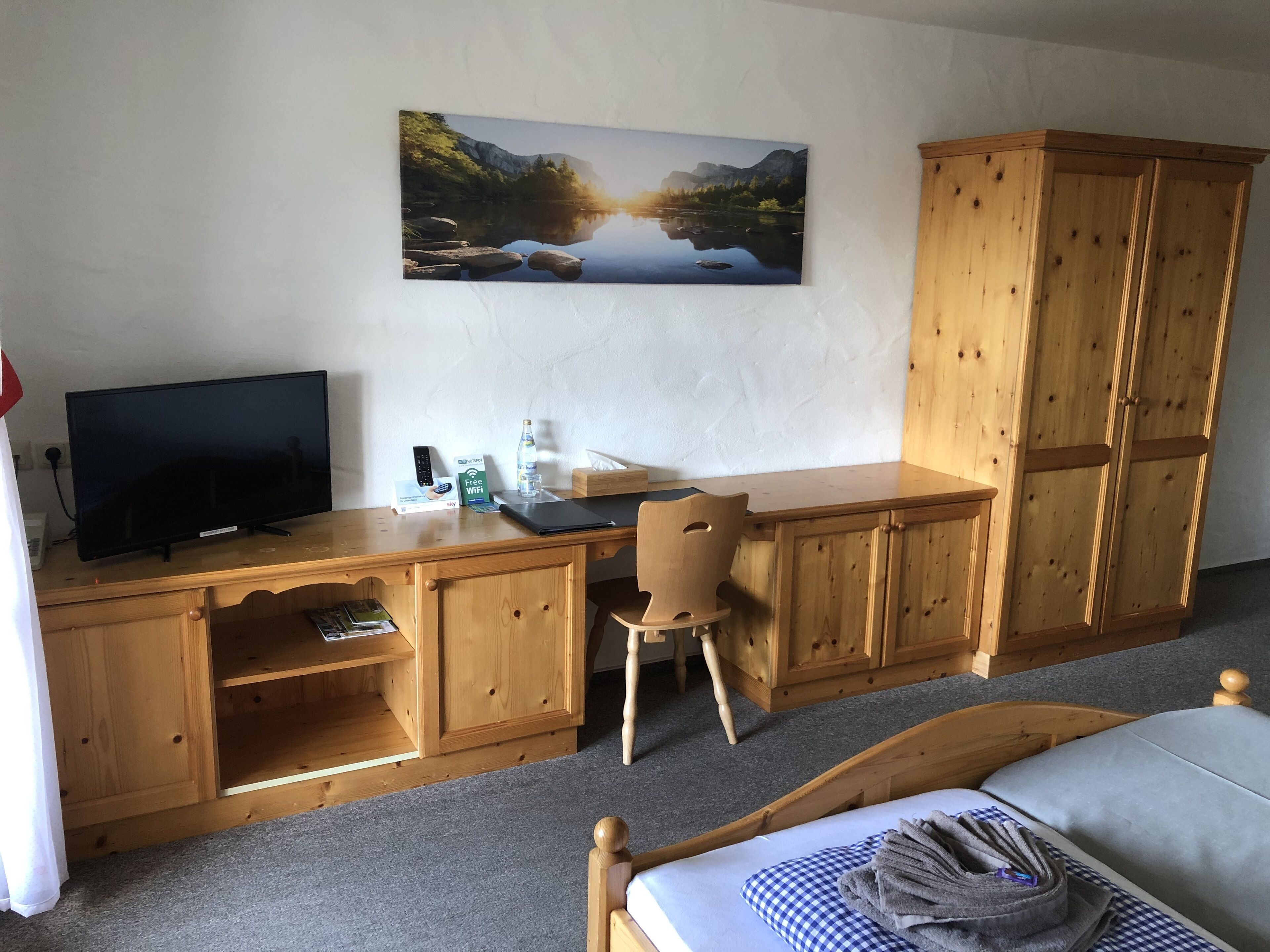 Une chambre comprenant un lit, un bureau avec une télévision, une chaise, une armoire en bois et un tableau représentant un paysage accroché au mur.