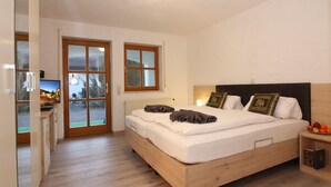 A bedroom with a wooden bed, two windows, a television, and a small bedside table with fruit. - PAGA Hotel (Aidenbach)