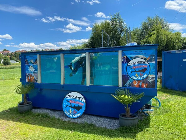 Un centre de plongée bleu avec des fenêtres en verre, portant les inscriptions « PAGA Diving » et « Tauch-Shop », entouré de pelouse et de plantes en pot.