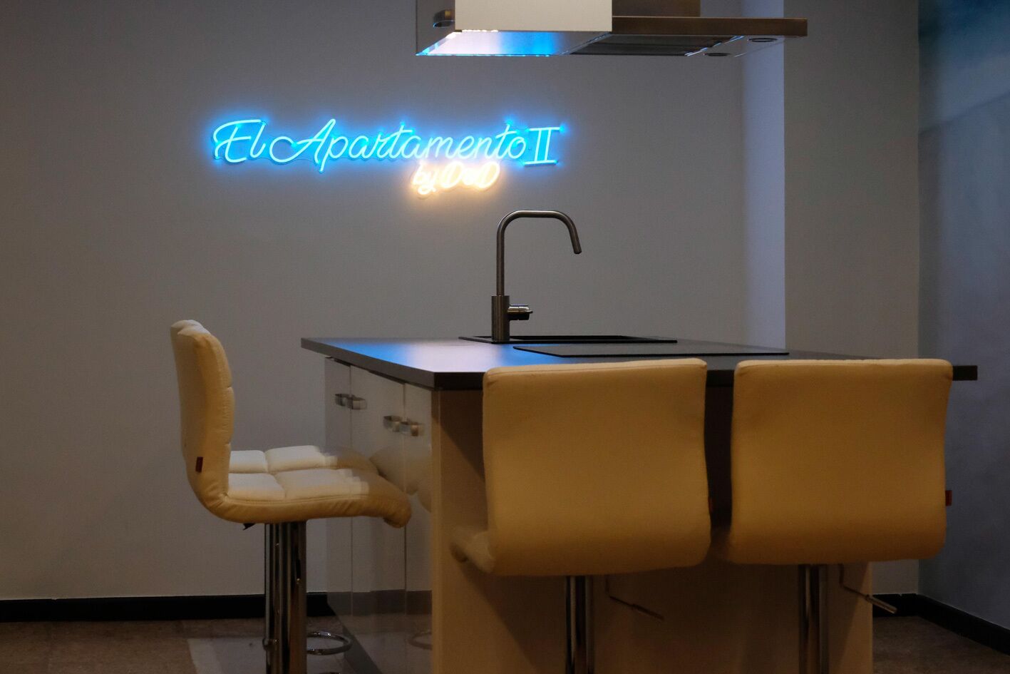 A modern kitchen with a neon sign reading "El Apartamento II" and a bar stool with a high backrest.