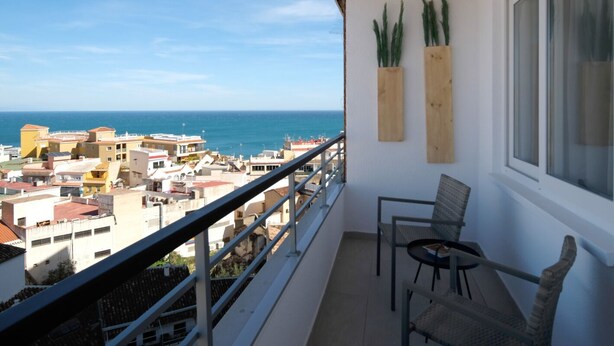 Un balcón con vistas al mar y edificios, una silla y dos macetas de madera.