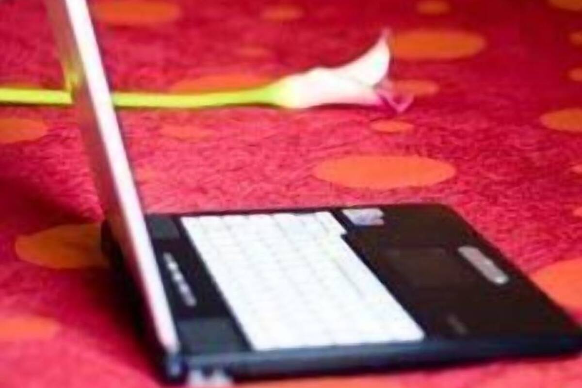 A laptop on a bed with a red polka dot bedspread, a bedside table with a lamp, and a wall-mounted light switch.