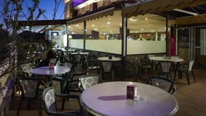 An outdoor seating area of a hotel with round tables and chairs, a neon sign reading "HOTEL L'ASTREHAZAS", and a view of the interior through the glass wall. - Hotel Las Terrazas (Albolote)