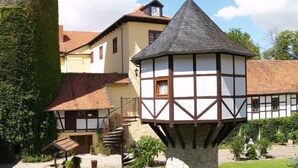 Une maison traditionnelle à colombages, dotée d’une tourelle, d’un puits en pierre et d’un jardin avec un chemin bordé d’alternances de pierres et de pavés.