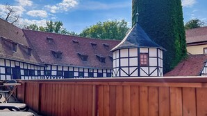 Une cour entourée d’une clôture en bois, une table avec une bouteille et un verre, et un bâtiment doté d’une tour et d’une tourelle.