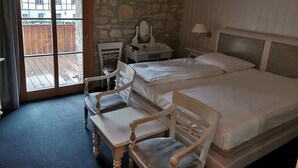 Une chambre d’hôtel avec un mur en pierre, un bureau en bois, une chaise, un lit et une vue sur l’extérieur, visible à travers une porte vitrée.