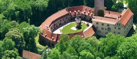 Vue aérienne d’un ensemble de bâtiments historiques comprenant une cour centrale, une tour de guet et une végétation luxuriante environnante.
