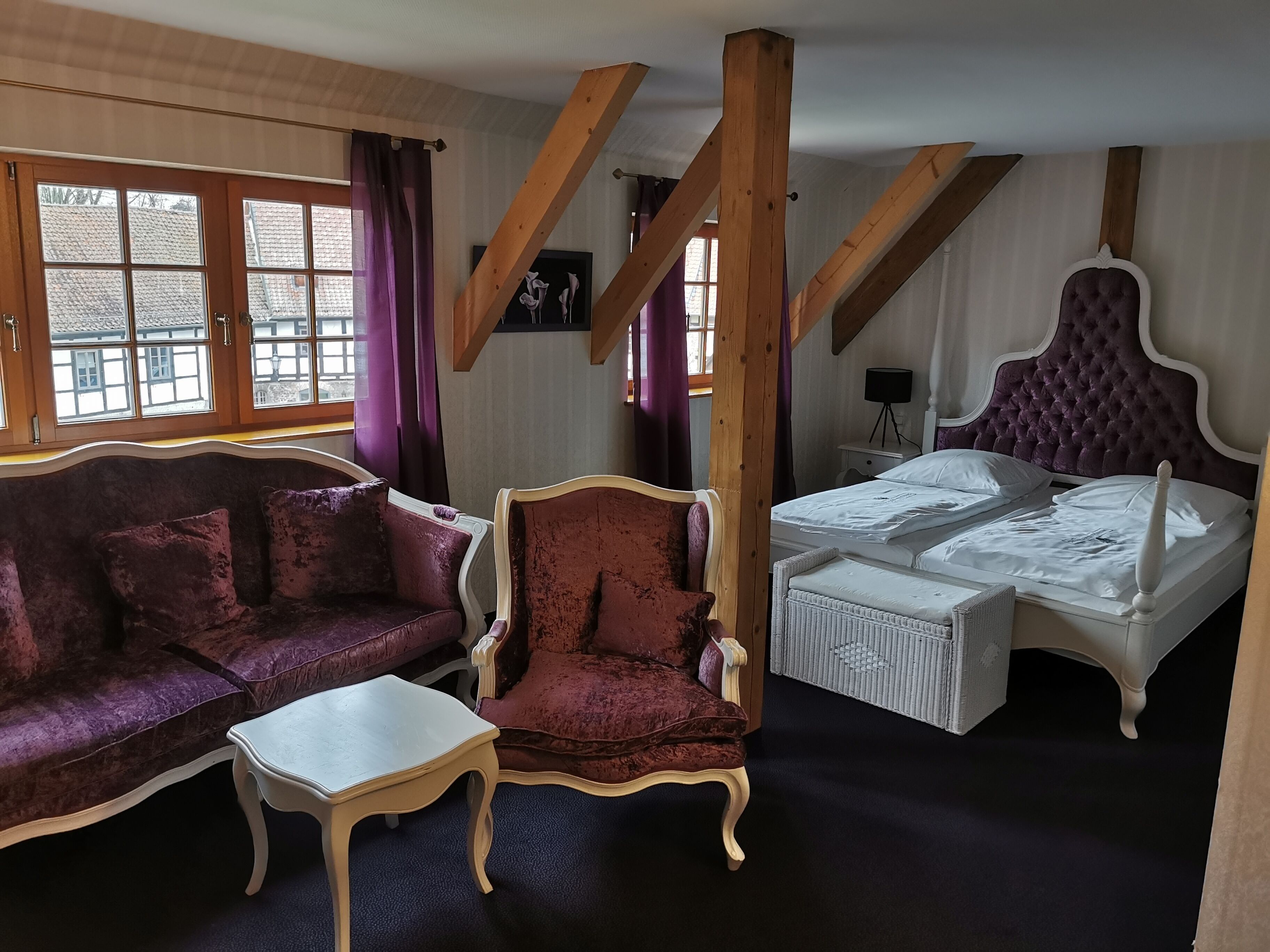 A room with a purple sofa, a matching armchair, a white bed, and exposed wooden beams.