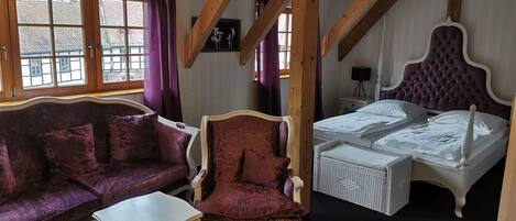 Une pièce avec un canapé violet, un fauteuil assorti, un lit blanc et des poutres en bois apparentes.