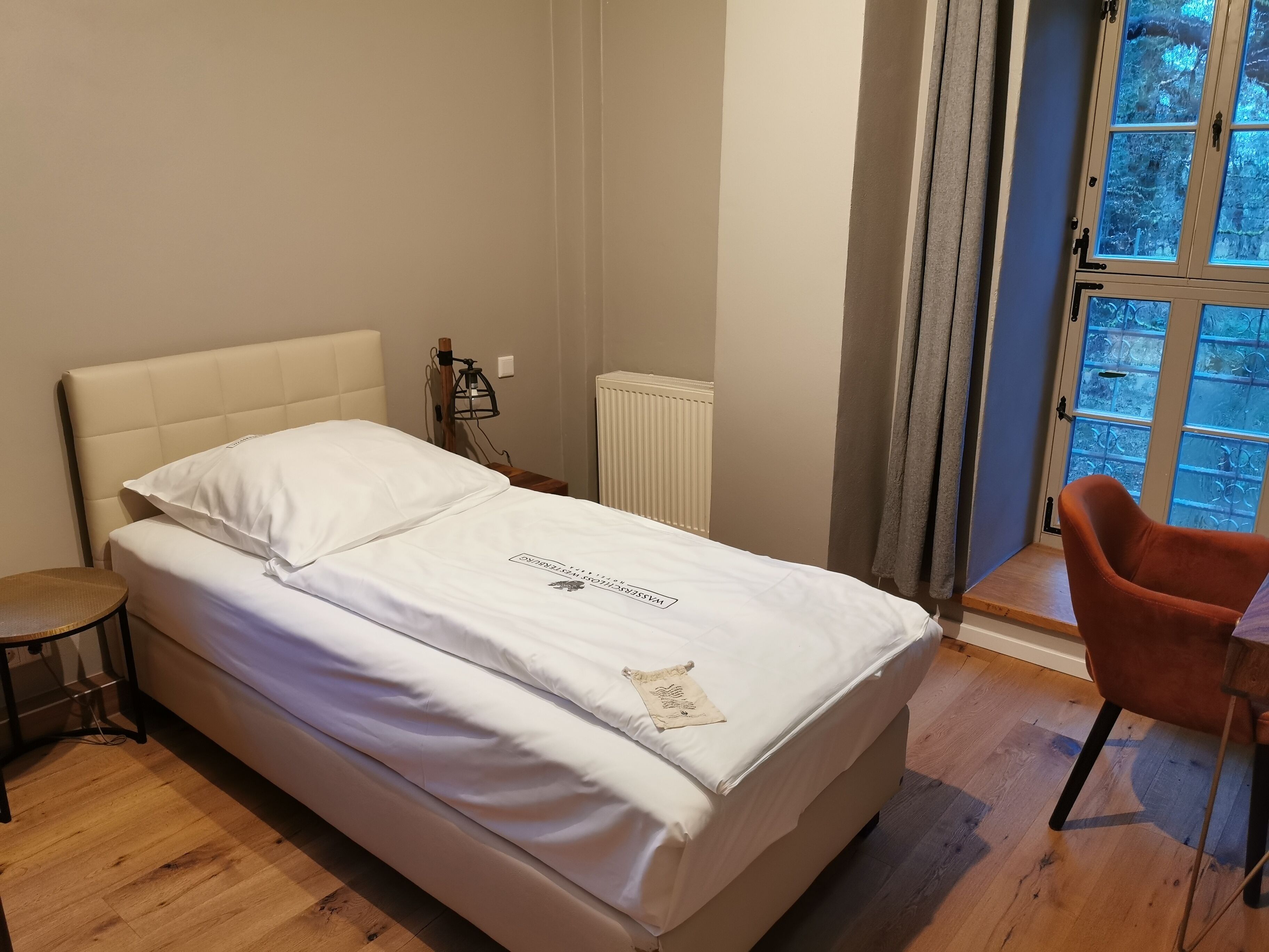 A neatly made bed with white linens, a bedside table with a lamp, and a chair.
