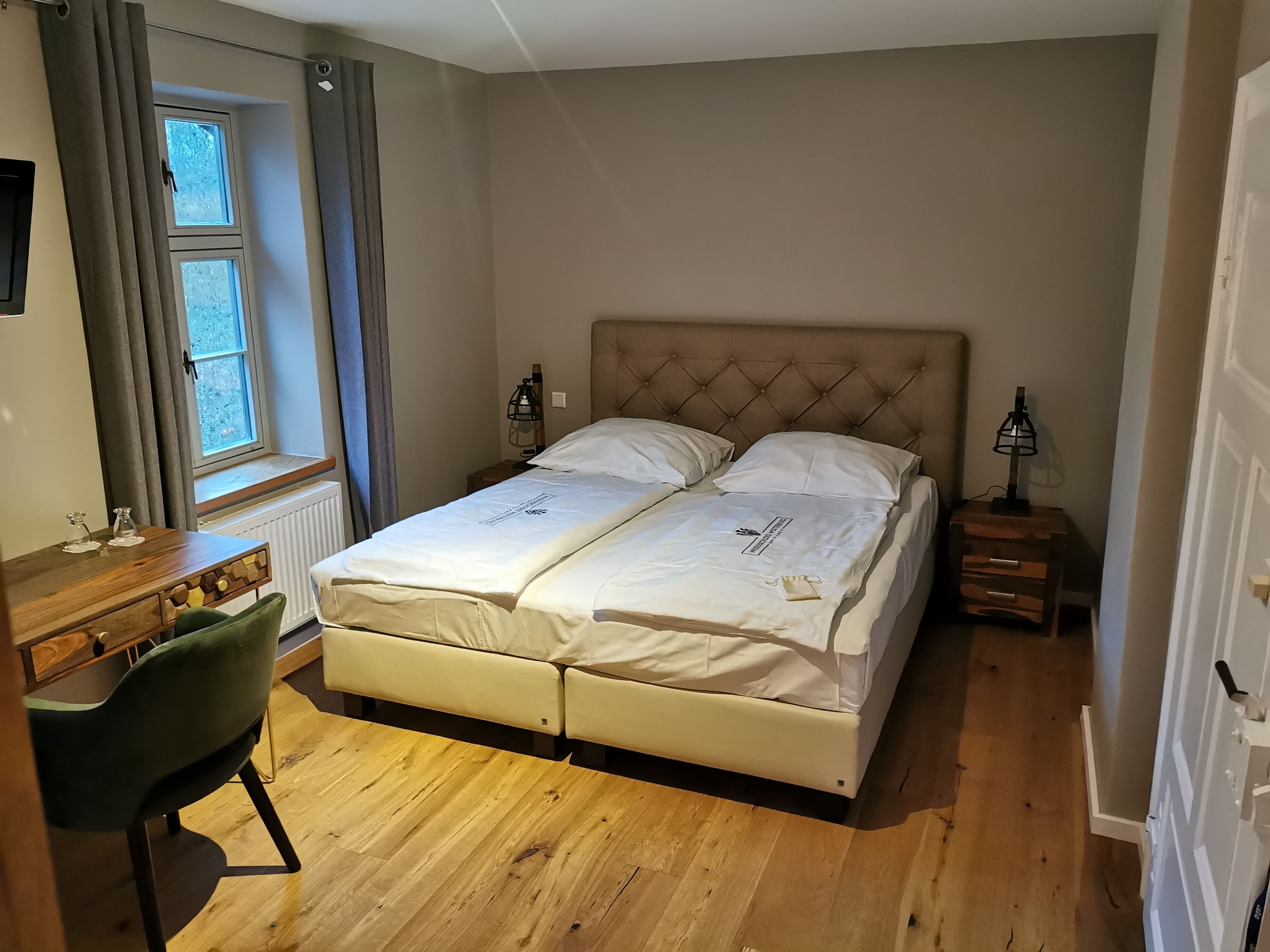A double bed with a tufted headboard, a bedside table with a lamp, a wooden desk with a chair, and a window with curtains.