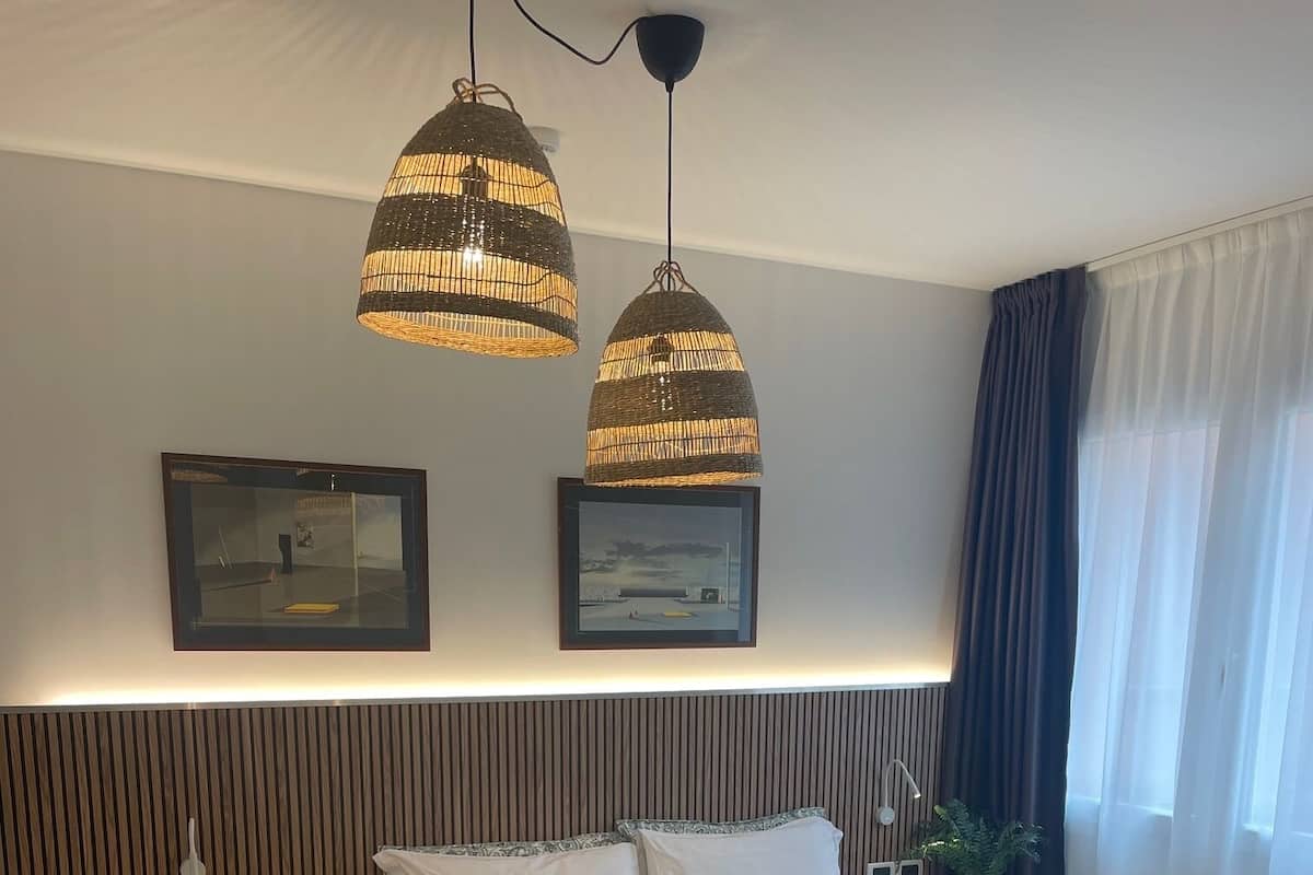 Une chambre d’hôtel moderne avec un grand lit, un canapé-lit plus petit, deux lampes suspendues, un téléviseur fixé au mur et une fenêtre avec des rideaux.