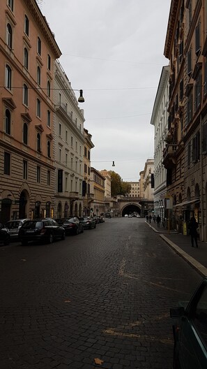 A cobblestone street lined with historic buildings, a pedestrian bridge, and parked cars.